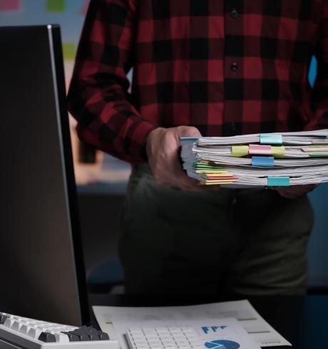 A man is holding a stack of papers in front of a computer monitor A man is holding a stack of papers in front of a computer monitor. The papers are organized and labeled with different colors